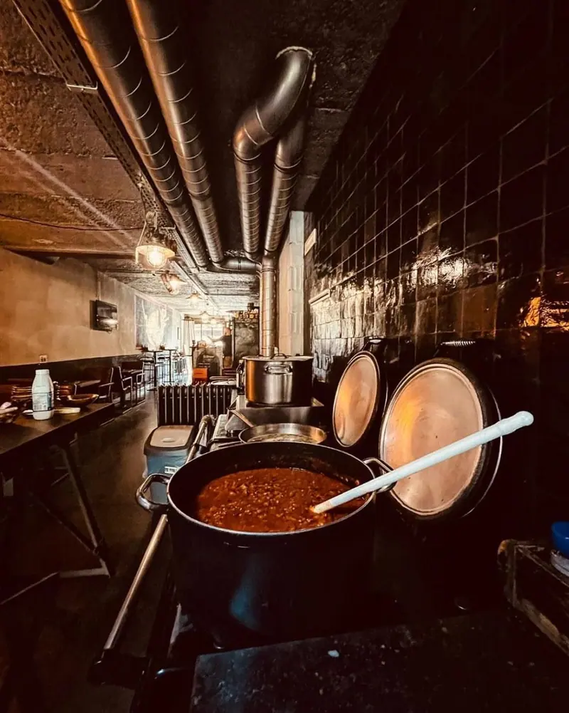 De open keuken van Kamiel in de avond: een pan met ragù op het fornuis, koperen deksels en stalen afzuigbuizen tegen een zwarte tegelwand.