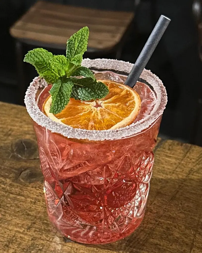 Een roze cocktail in een kristallen glas met een gesuikerde rand, een schijfje sinaasappel en munt.