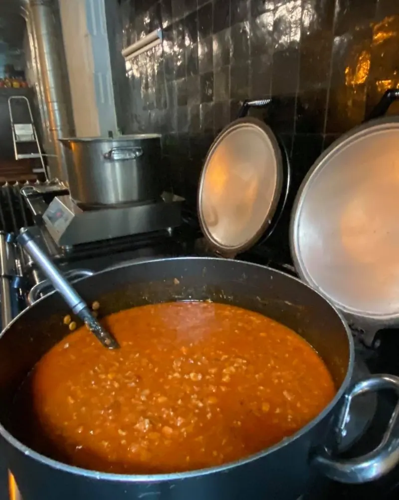Een grote pot ragù op het fornuis, met koperen deksels en stalen kasten erachter.
