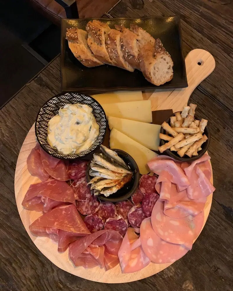 Een ronde plank met ham, salami, kaas, sardientjes, brood en een dip.